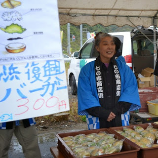 「浜復興バーガー300円」と書かれた手作りの看板が掲げられ、七の市商店街の法被を着た女性が笑顔でバーガーを販売している様子の写真