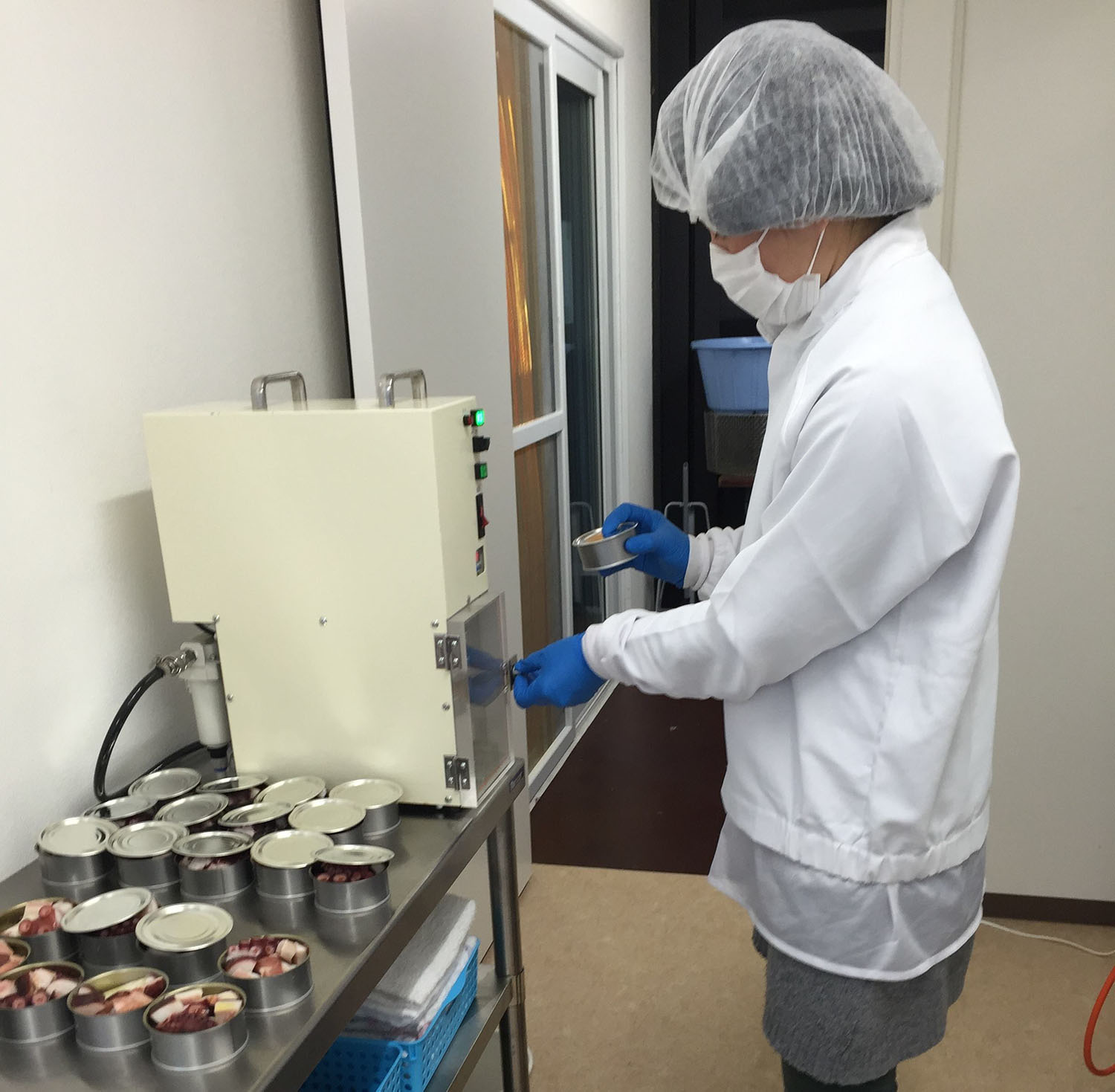 白衣と衛生帽、マスクを身につけた人物が、ステンレス台に並べられた缶詰の中身を機械で封をしている作業の様子の写真