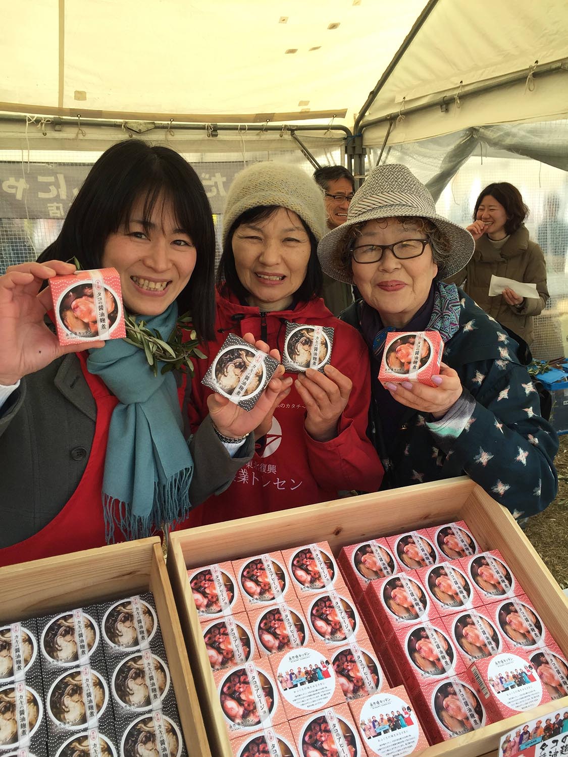 木箱に缶詰がたくさん陳列されているテントの下で、中村悦子さんと2人の女性が缶詰商品を手に持ち笑顔で並んでいる写真