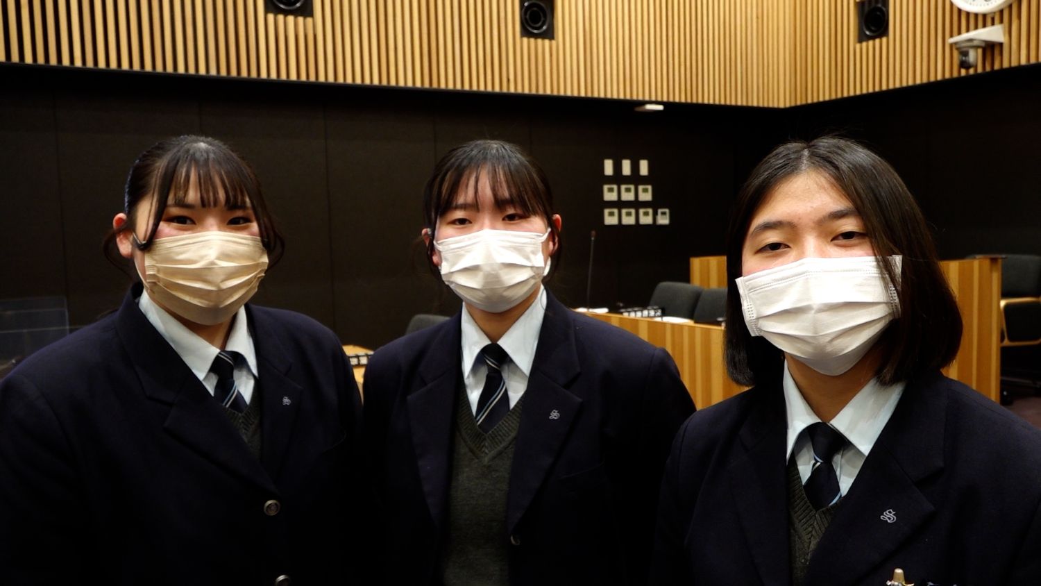 議場内で、紺色の制服にネクタイを締めた女子高校生3人がマスクを着けて並び、カメラに向かってまっすぐ立っている高校生の集合写真
