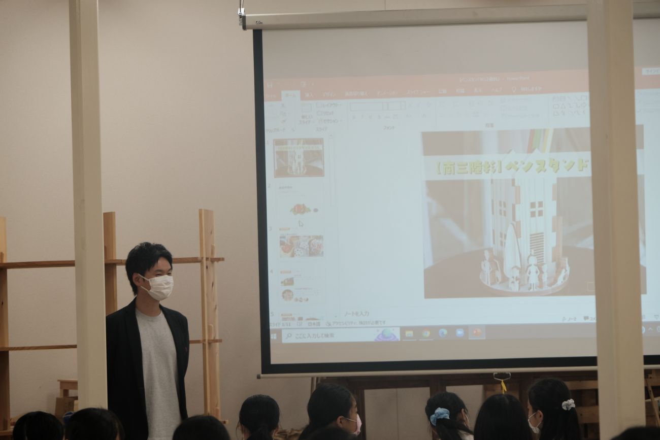 プロジェクターで木製のペンスタンド模型の完成見本を映し出しながら、マスクを着けた大森丈広さんが前に立ち児童たちが説明を聞いている教室の様子の写真