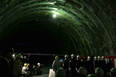 一か所だけ灯りが灯された暗いトンネル内にヘルメットを着用した人々が集まっており、代表者が横一列に整列して立ち、左側に座っている人たちがカメラを構えている貫通式の様子の写真