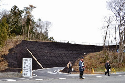 傾斜地に黒い法面保護材を施した完成した道路に、「竣工式並びに団地見学会会場」と書かれた看板が設置された入り口に警備員が配置され、スーツ姿の2人が入り口から出てきて歩いている写真