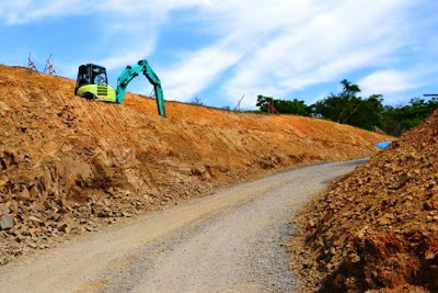 両側を削られた赤土の中央をカーブする未舗装の造成道路と、傾斜地で作業を行っている重機が見える写真