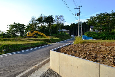 舗装された新しい道路の脇に工事中の重機と、奥に住宅地が見える静かな風景の写真