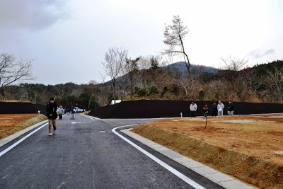 土地の造成が終わったばかりの広い舗装道路に人々が歩いており、両側の斜面が黒い土で固められている分譲地の写真