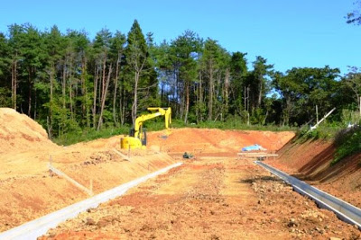 赤土を広く掘り下げた土地で黄色い重機が道路の基礎を整備している写真