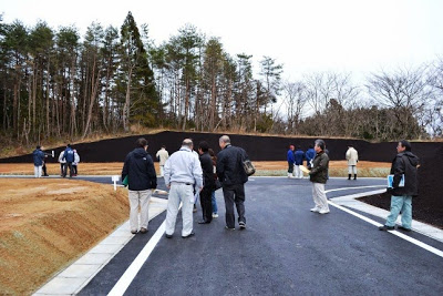 森の傍で黒い法面と舗装道路が完成し、分譲地を多くの人が見学している写真