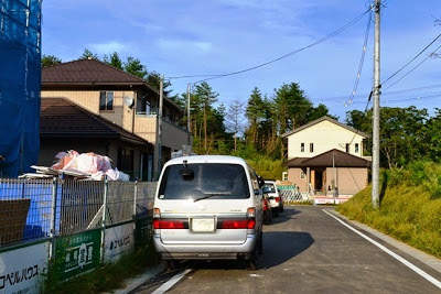 新築住宅が立ち並ぶ住宅地の細い舗装道路に白いワゴン車が停まっており、左側には建築資材が積まれたブルーシートで覆われた建設現場がある新興住宅街の写真