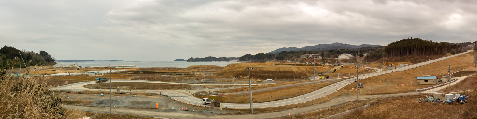 画面には曲がりくねった道路が複数あり、建設車両や機材が点在し、草地やいくつかの建物が含まれ、背景には丘が広がり画像の左側には海が見え、遠くには水面に突き出た陸地が確認できる海沿いの地域の写真