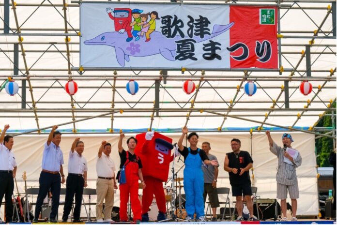 舞台の上で複数の大人が並び、中央に赤いキャラクターの着ぐるみがあり、背景に「歌津夏まつり」と書かれた横断幕が掲げられている、祭りの開会式の写真