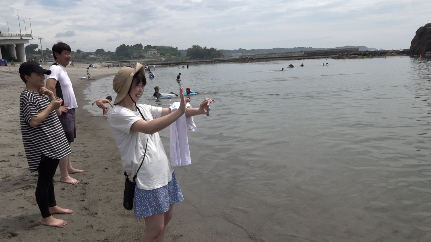 後ろには楽しそうに様子を見守る大人たちや子どもが見える砂浜の水際で帽子をかぶった女性がスマートフォンで海に入っている人たちを笑顔で撮影している写真