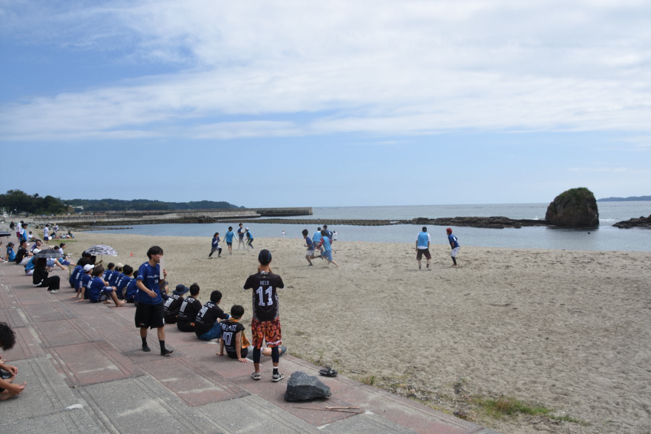 ビーチでアルティメットの試合が行われており、観客が座って試合を見守る中、紺色のユニフォームのチームと水色のビブスのチームが砂浜で競技をしている様子の写真