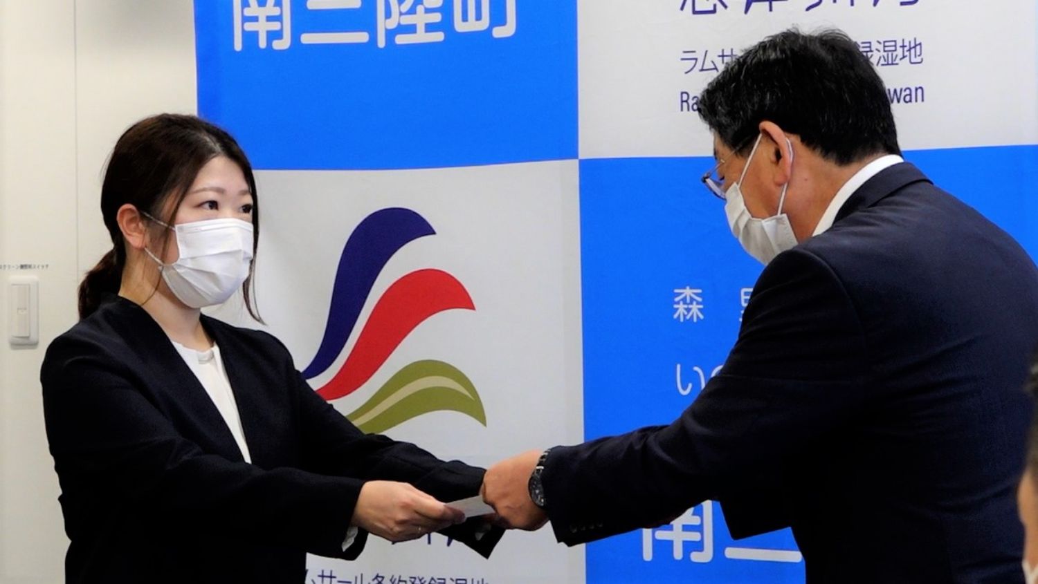 南三陸町のバックパネルの前で、スーツ姿の太田さんがマスクをして佐藤仁町長から委嘱状を受け取る様子の写真