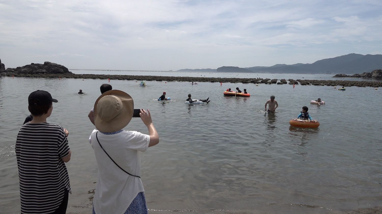 帽子をかぶった女性とボーダーの服の女性が海辺からスマートフォンで波の穏やかな浅瀬で浮き輪に乗った子どもや楽しそうに水遊びをする大人を撮影している写真