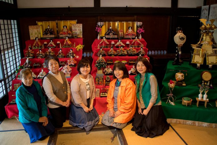 7段飾りの豪華な雛飾り2つと3段の兜飾りの前に、5人の女性が並んで座っている写真