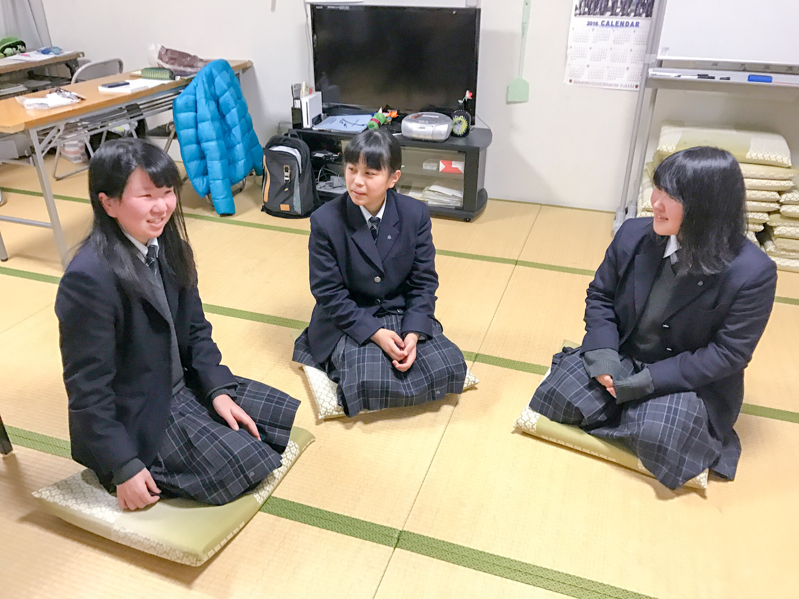 テレビや長机が置かれた和室の部屋で、座布団の上に座って話をしている制服姿の女子高校生3人の写真