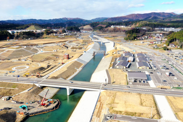 川沿いに建設が進む防潮堤や道路、商業施設の建物群と駐車場が写っており、奥には山々と街並みが広がっている写真
