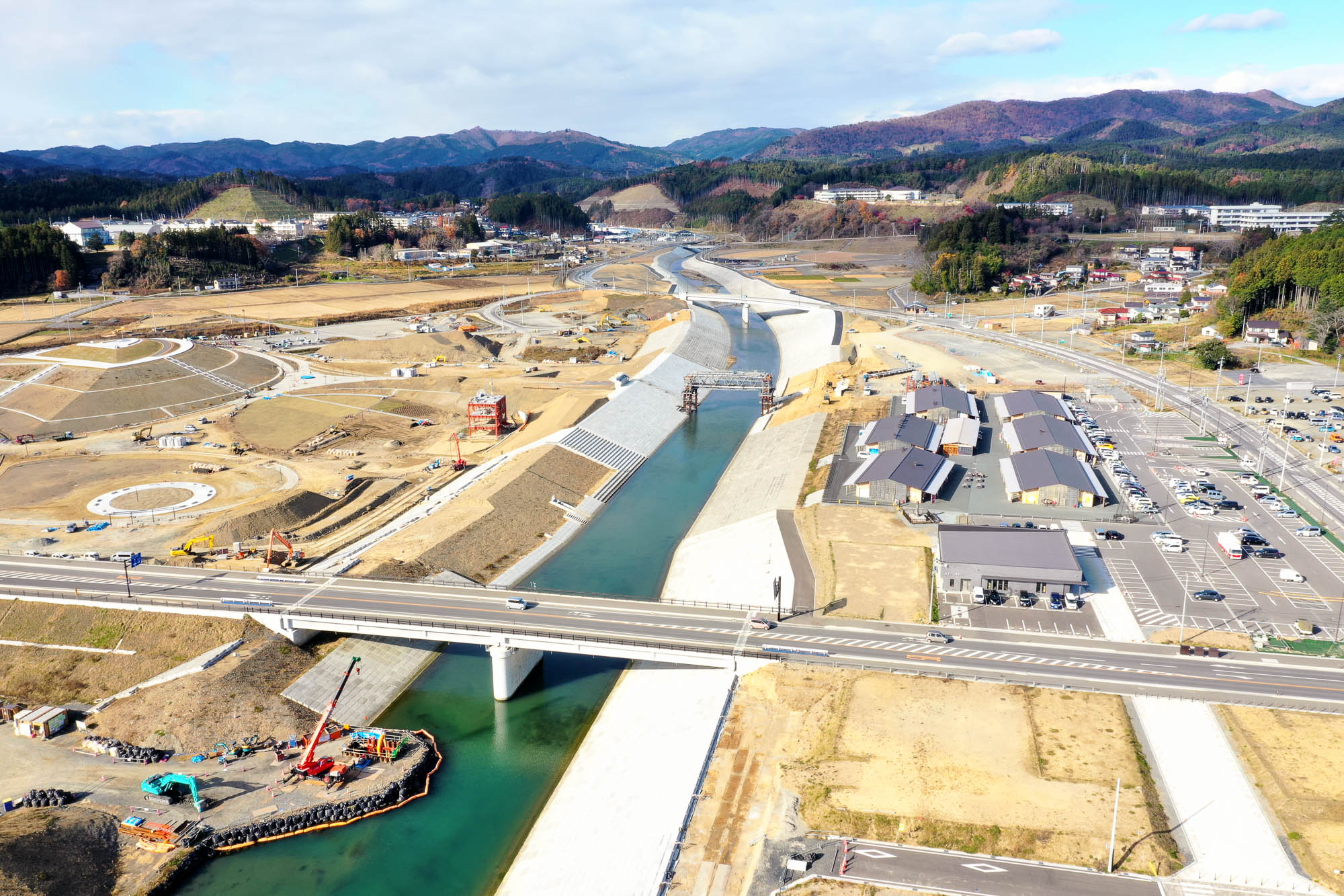 中央を川がまっすぐ流れ、左右にコンクリートの護岸が整備され、手前に橋、右側に仮設商店街や駐車場、奥に山々が見える様子を真上から撮影した写真