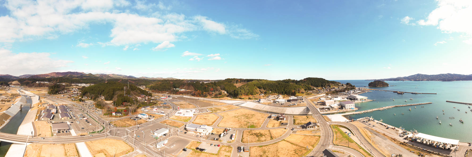 手前から奥にかけて河川が流れ、山々に囲まれた造成地の広がる風景と、右手に住宅地や商業施設が並び、さらに奥に海が広がる様子を広角で写した写真