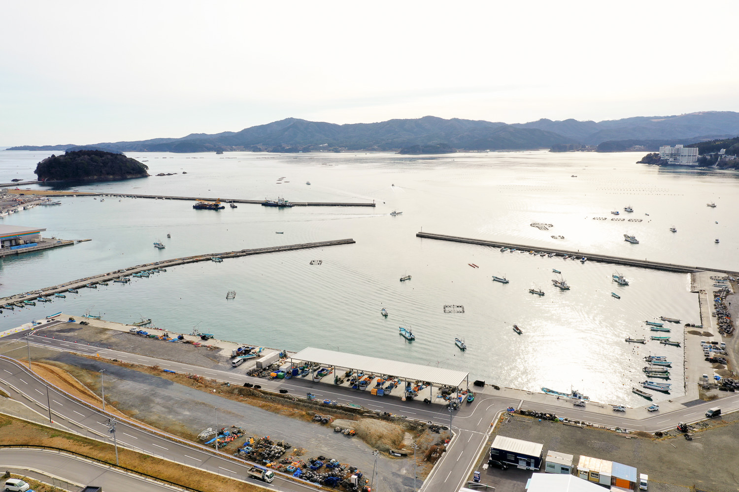 防波堤に囲まれた港に多数の小型船が停泊しており、海岸沿いには倉庫のような建物が並び、左側には島が見え、背景には丘や山々が広がる穏やかな海辺の風景の写真