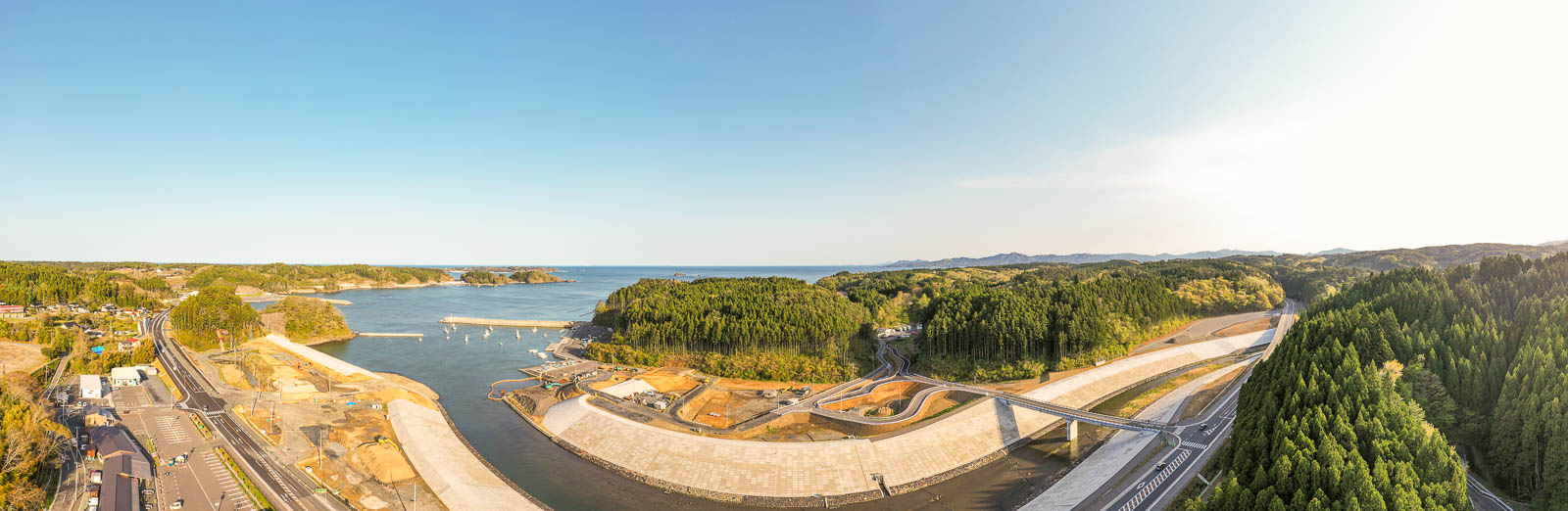 中央に海へと注ぐ川とその両岸に建設された大規模な護岸構造物、右側に深い緑の森林と新しく架けられた橋、左側に沿岸の街並みと道路が見え、遠くに小さな港と島々が広がる様子を快晴のもとでパノラマ撮影した沿岸地域の復興風景の写真