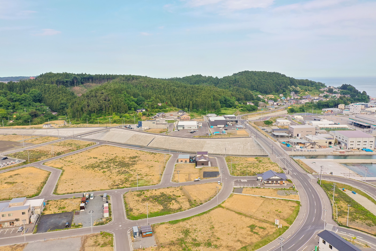 住宅や商業施設が混在する沿岸の町を空から見下ろし、交差する道路、空き地、緑地が点在し、背景には森林に覆われた丘と右側に海岸線が広がる写真