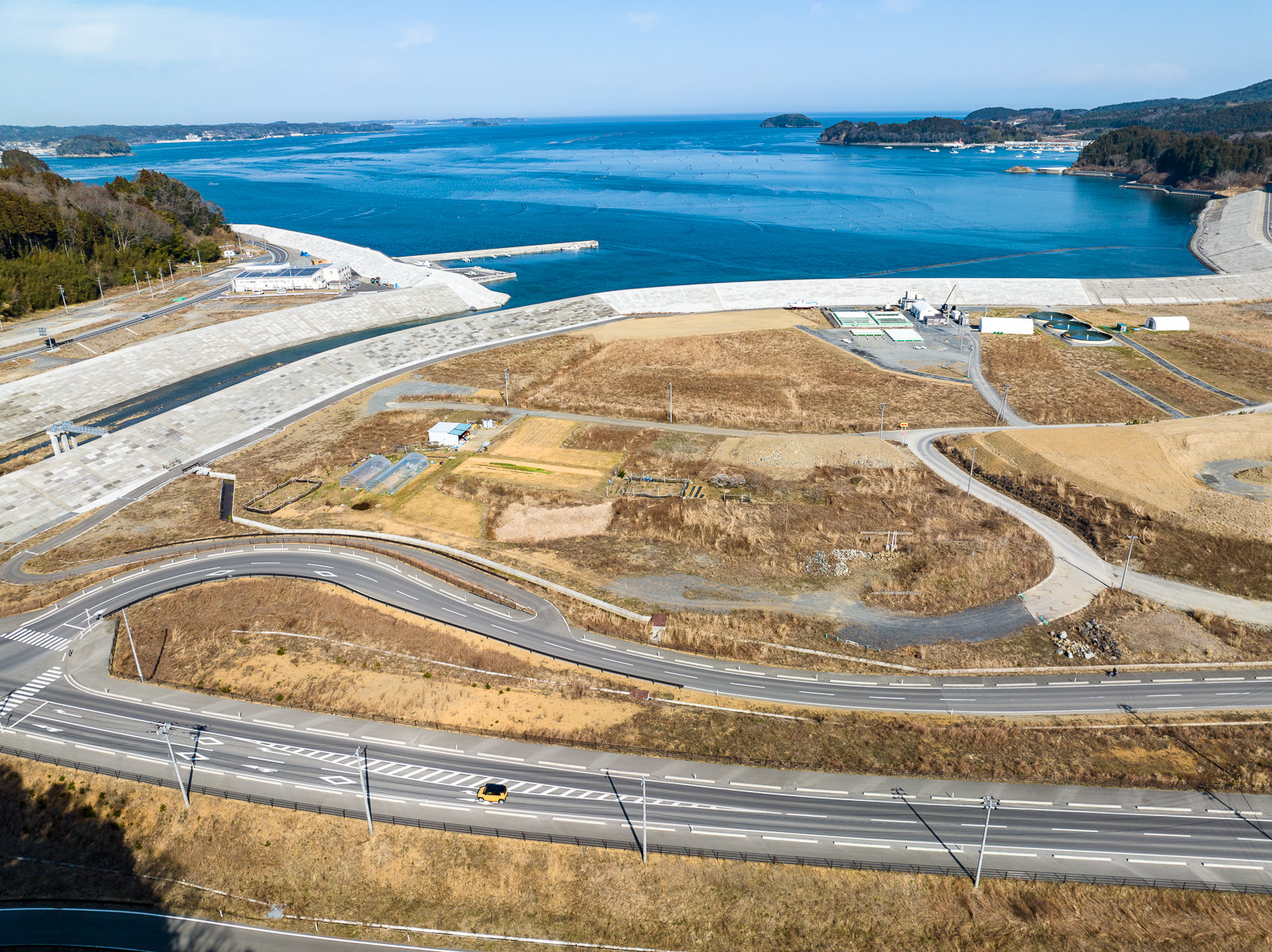 複数の防潮堤が重なるように続き、奥には静かな湾と島が見え、手前の道路はカーブしながら土地の起伏に沿って走っている、海辺の復興エリアの昼間の風景を高台から定点カメラで撮影した写真