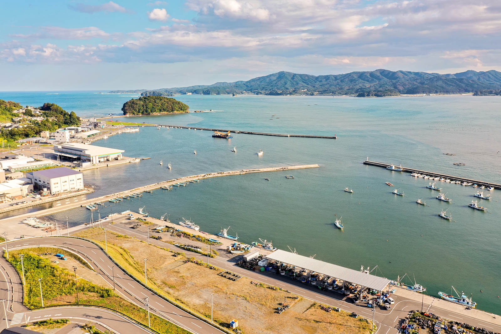 手前に防波堤と漁港の施設群、その向こうに漁船が浮かぶ港、左奥に森に覆われた小島が見える湾西側の写真