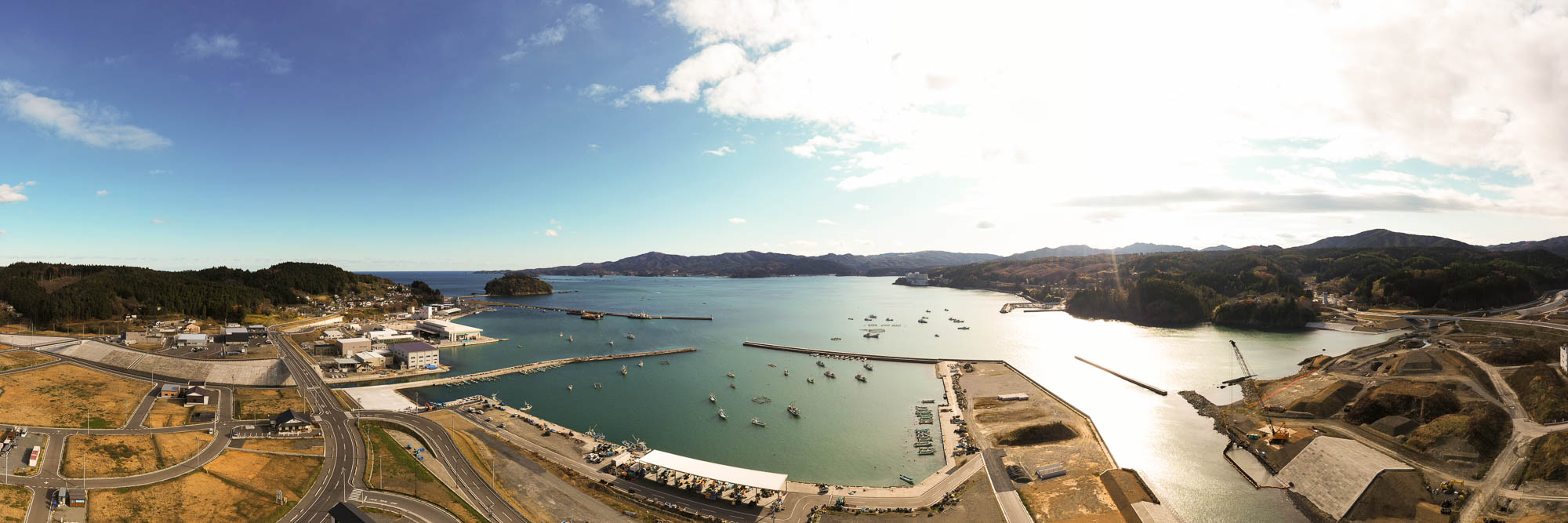 港全体と山々、町の様子が一望できる空撮で、左側には港町と林に囲まれた住宅地、右側には工事中の斜面や道路が広がっている港と町の全景の写真