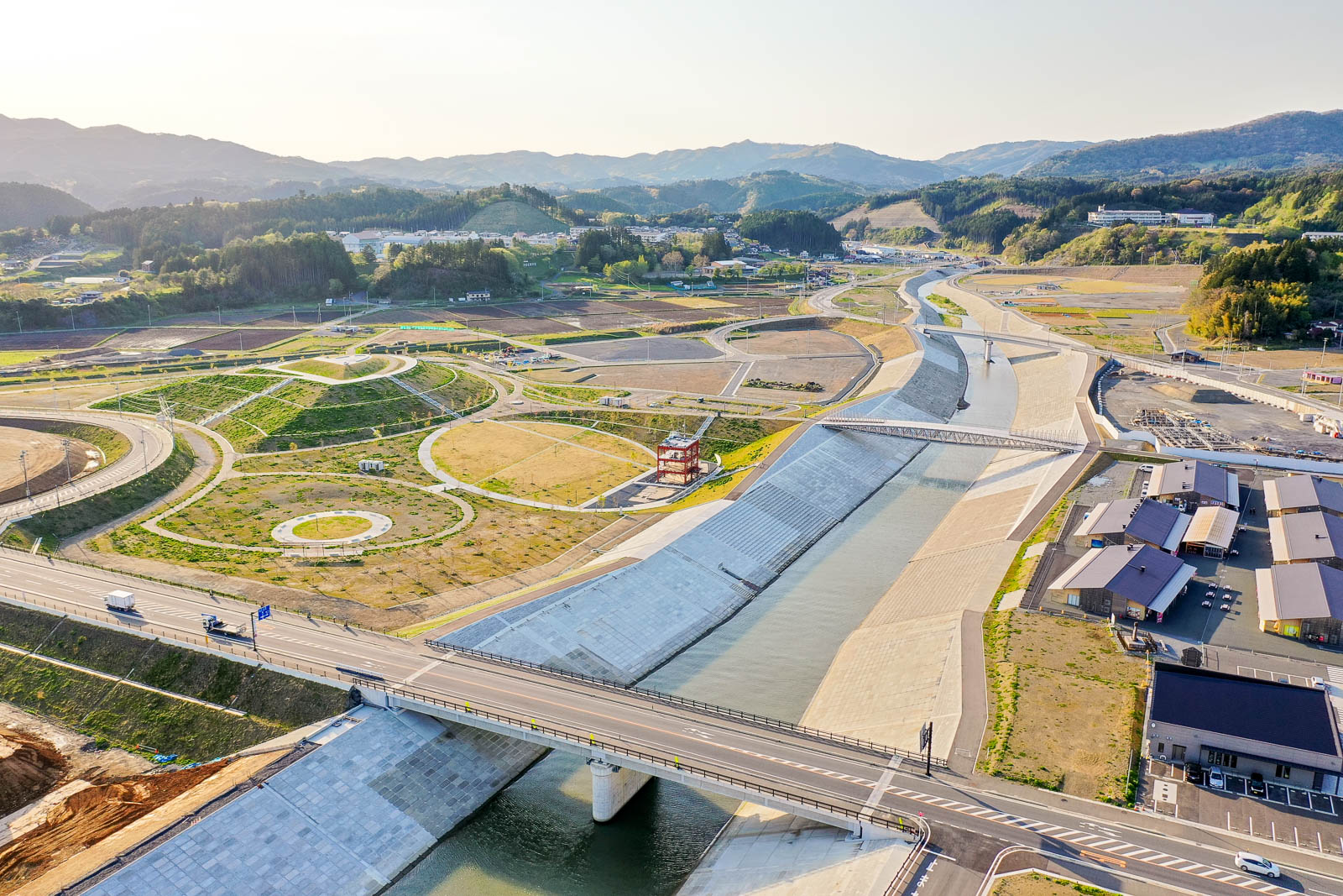 中央にはコンクリートでしっかり護岸された川が流れており、堤防の上には車道と歩道橋が設けられていて川の左側には円形や曲線を多用した広場や緑地が整備され、公園のような空間になっている写真