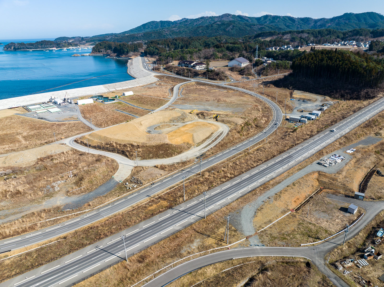 舗装された道路が蛇行しながら丘を登り、右手に造成中の土地区画や重機が見え、奥には湾と山が広がる、再開発中の沿岸地域の昼間の風景を高台から定点カメラで撮影した写真