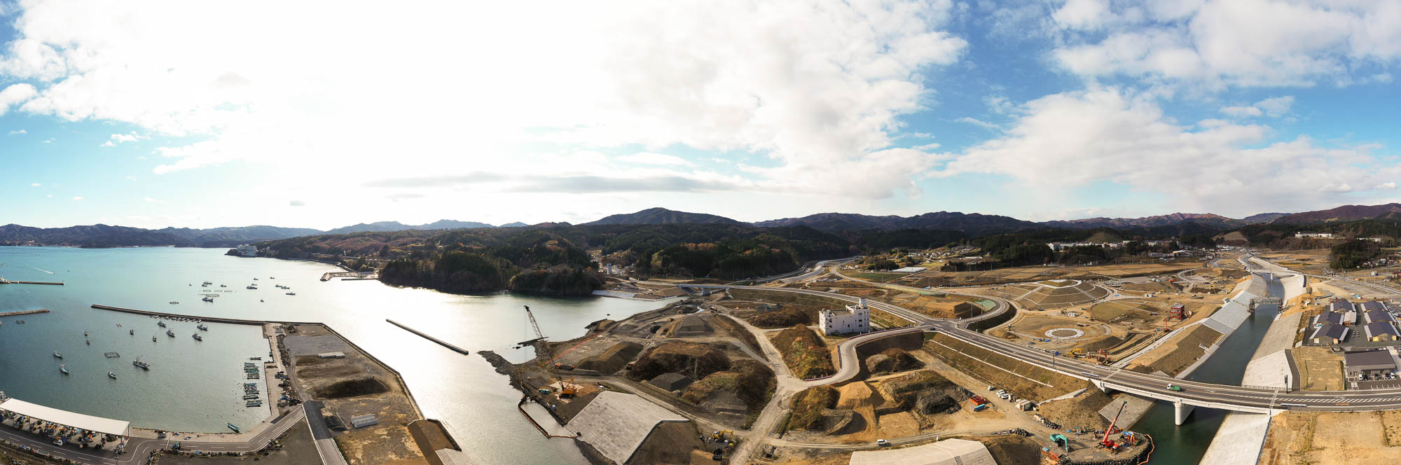 海に面した工事現場とその奥に広がる道路網や住宅地が見え、山に囲まれた町の風景と整備中の土地が広がっている海と山と造成地の写真