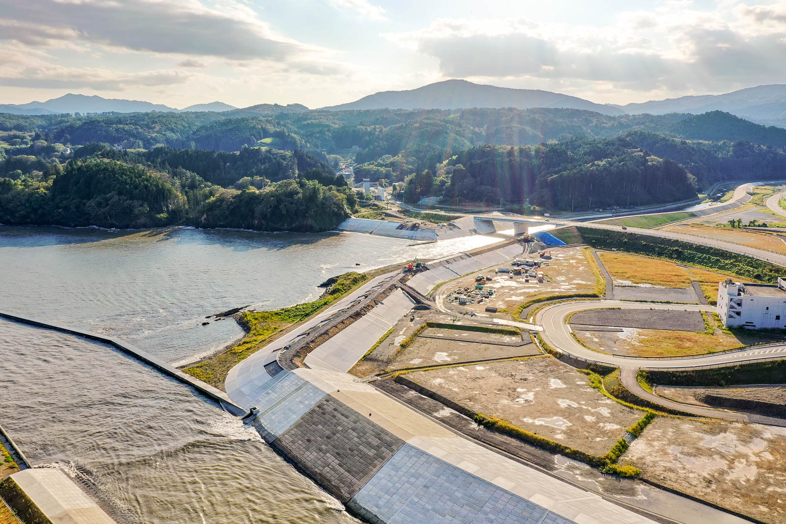 川沿いに建設中の護岸と作業車両、その背後に続く緑の山々と住宅地、手前に茶色い更地が広がる市内陸部の写真