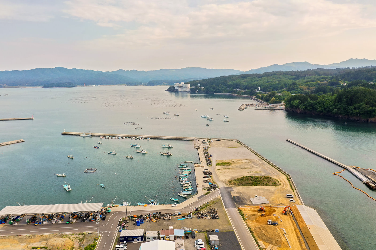 港に複数の船が停泊し、周囲に建物や道路、空き地が広がり、背景には丘と山々が連なり、穏やかな海と曇りがちの空が見える沿岸地域の写真