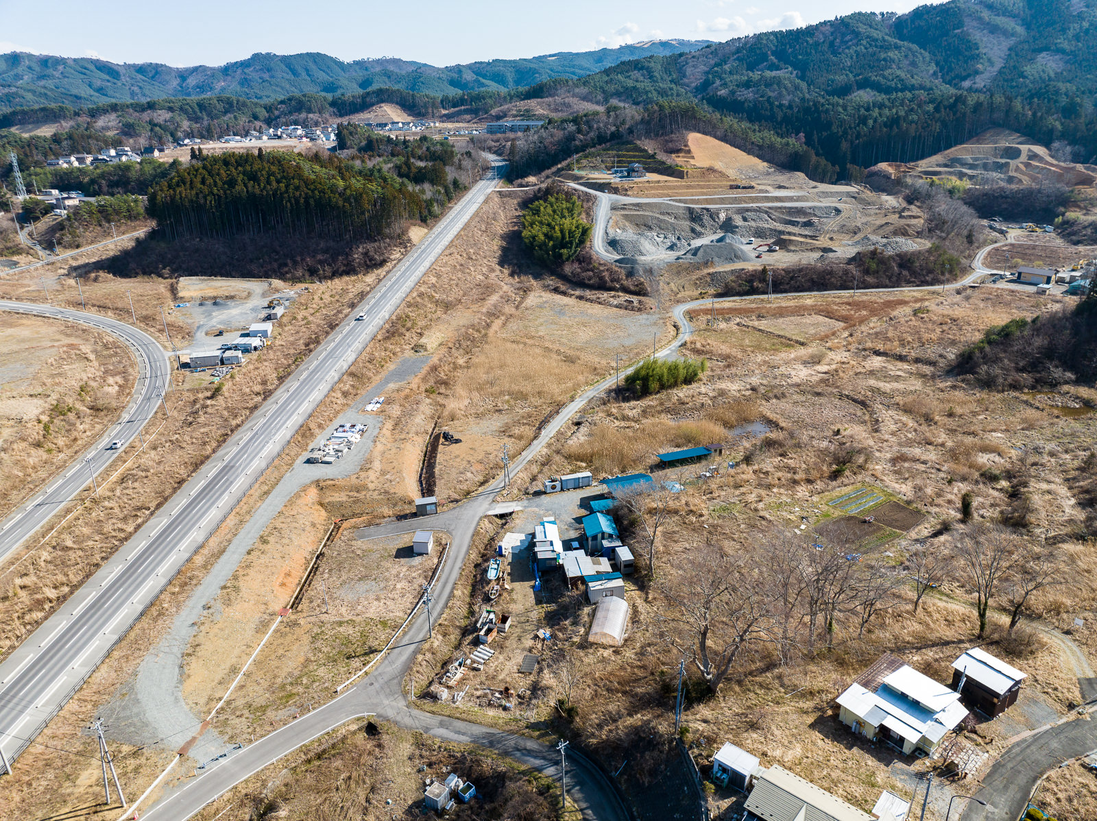 山間部の麓から高台へ伸びる幹線道路沿いに、資材置き場や仮設建物、小規模な作業場が点在しており、遠くには住宅地が広がる、復興インフラの終点部の昼間の風景を高台から定点カメラで撮影した写真