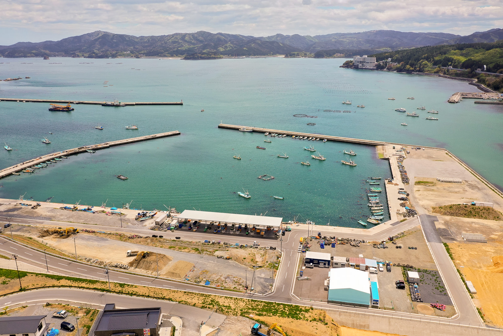 青く澄んだ海に複数の桟橋が伸び、保護された港内に多くの船が停泊し、周囲には建物や駐車場、道路が広がり、背景には緑に覆われた山々が見える海辺の町の写真