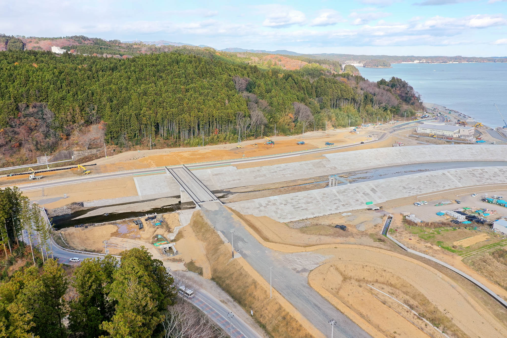 森に囲まれた中を流れる川に架かる橋と、その周囲で整備が進む造成地、奥に広がる海岸線が見える写真