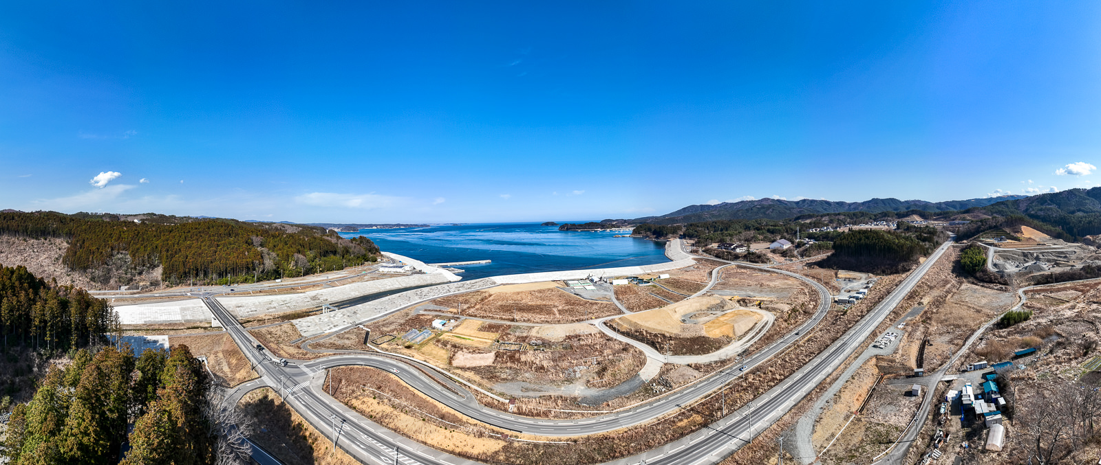 奥には海と防潮堤、手前には新しい道路が曲線を描いて走る風景が広がっている、海岸部の再整備中の昼間の風景を高台から定点カメラで撮影した写真