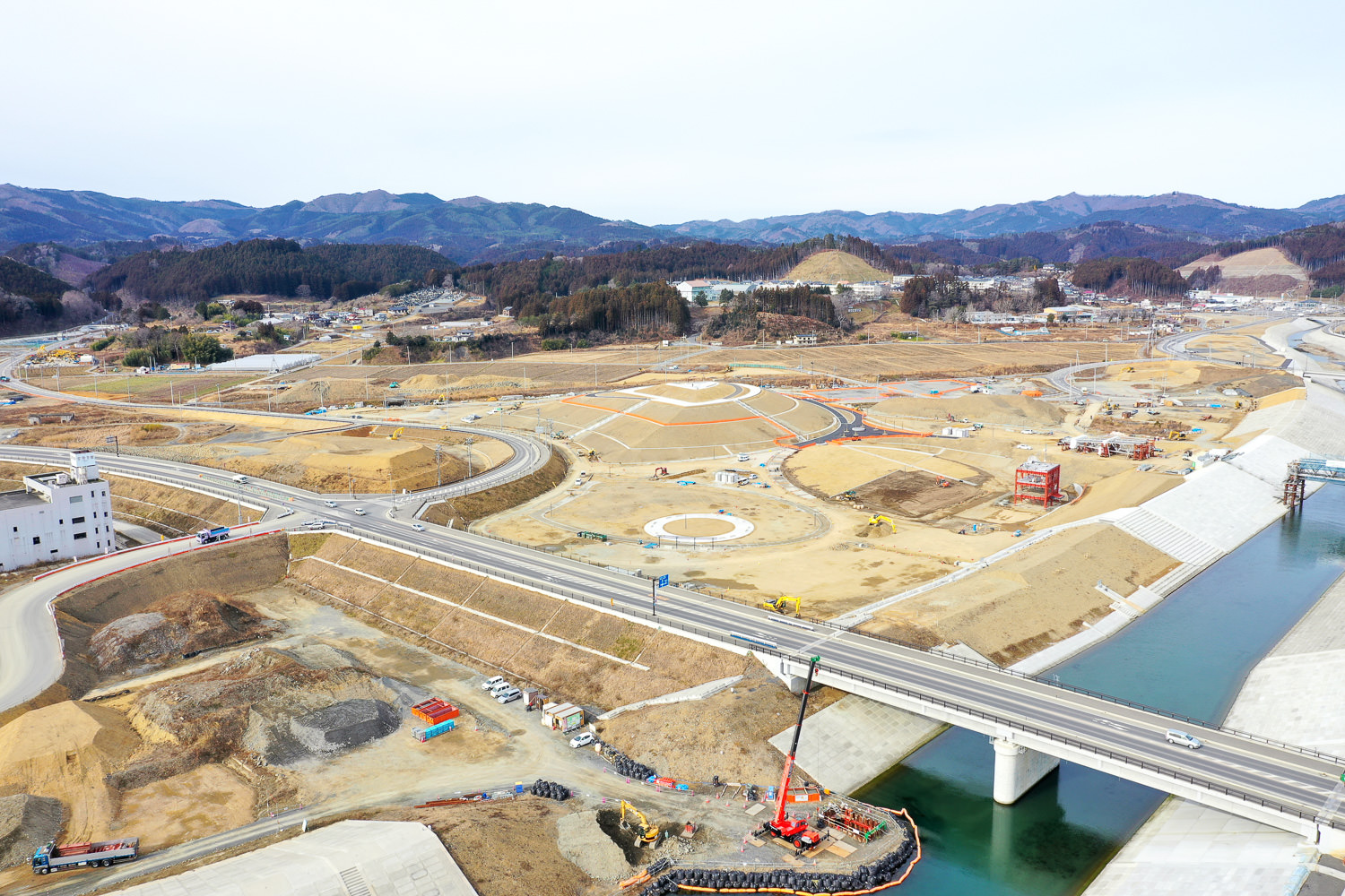 川に架かる橋の手前に広がる大規模な工事現場では、複数の円形の基礎構造や道路が建設中で、建設車両や機材が点在し、背景には丘や山々が広がっている工事現場の写真