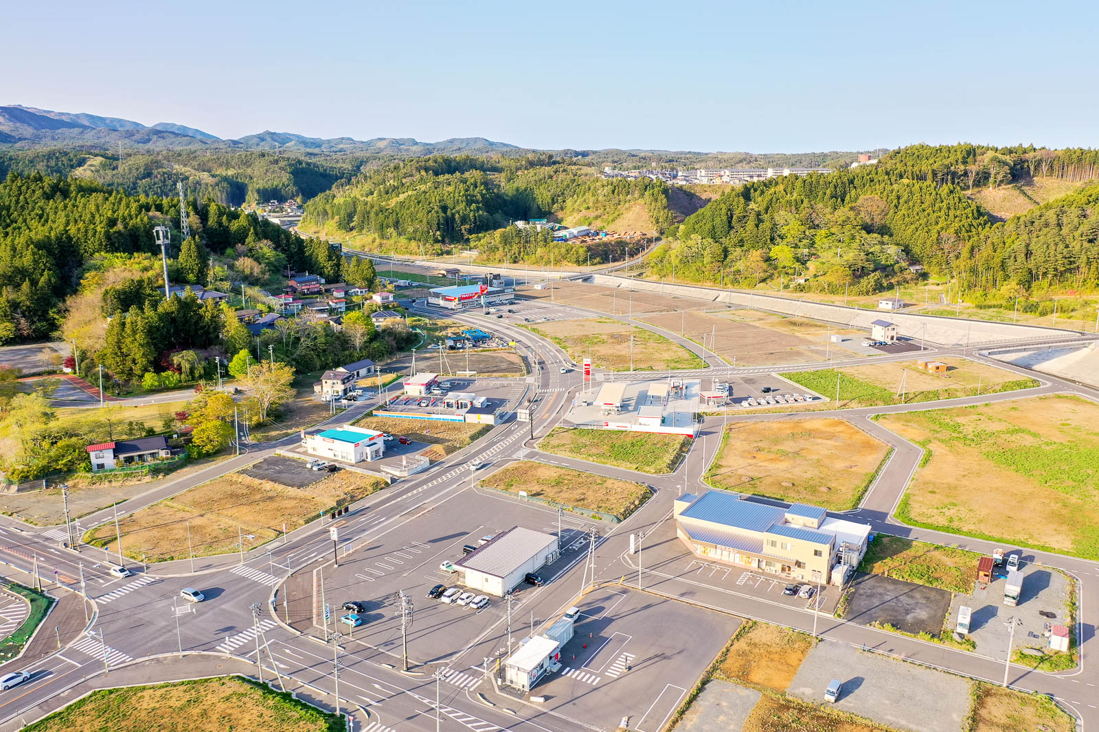 手前には広い交差点と整備された駐車場付きの建物があり、その周囲は区画整理された空き地や新しい道路が広がっている写真