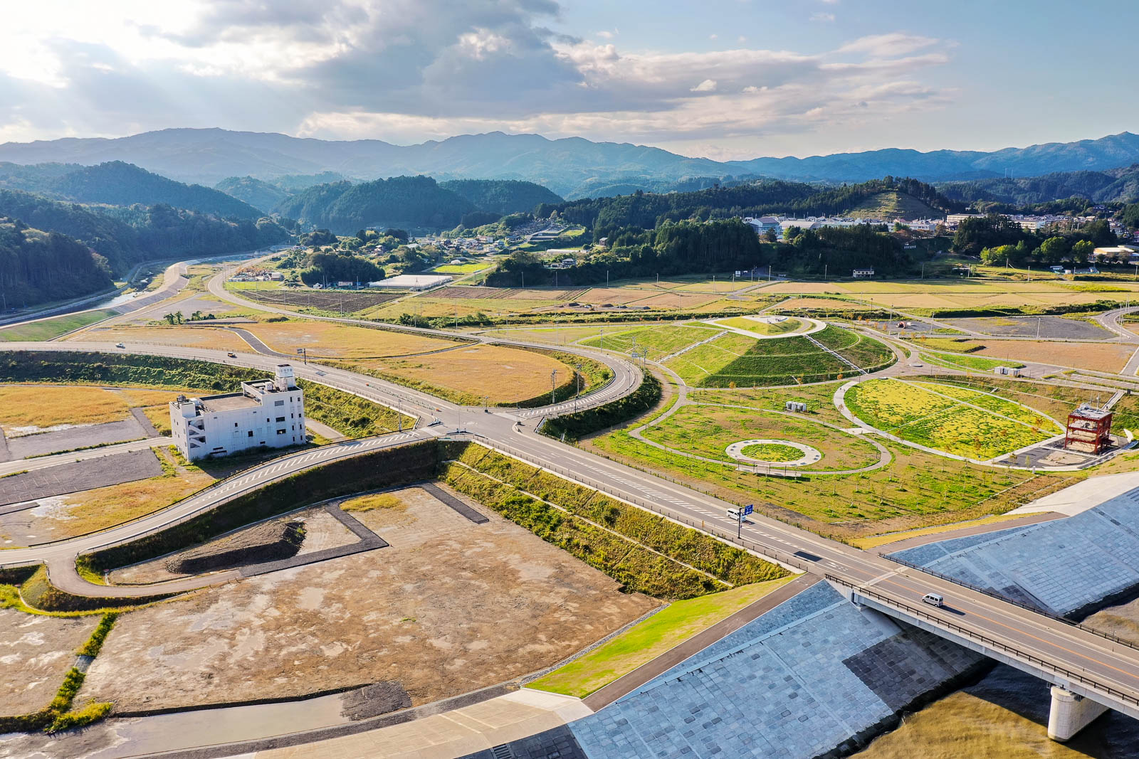 中央の円形広場を囲む緑の芝生と道路網、背後に広がる田園と山々が見える市の復興造成地の写真