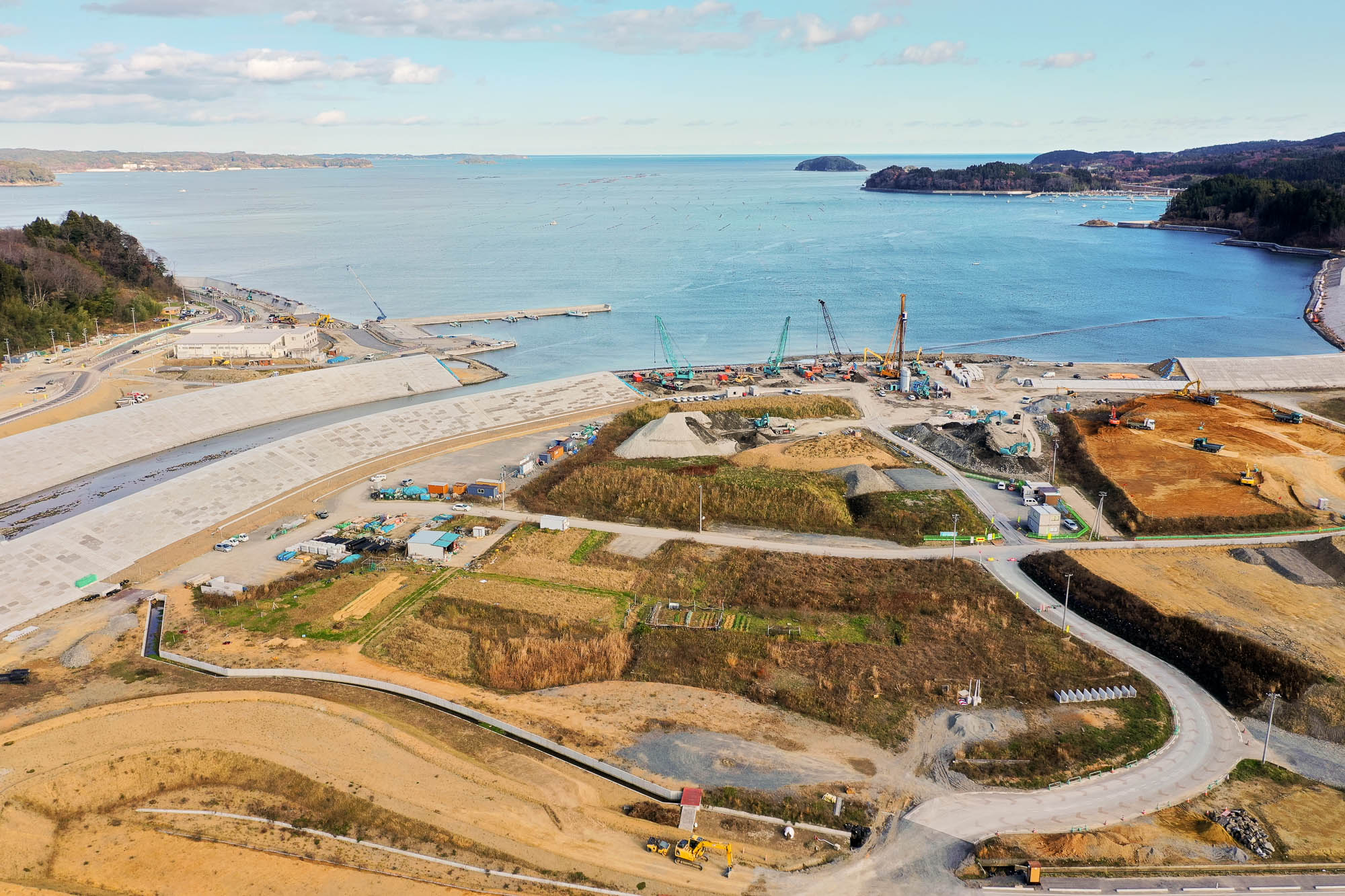 防潮堤の奥に広がる港湾エリアで大型機材やクレーンが並び、湾内には防波堤や漁港が見える写真