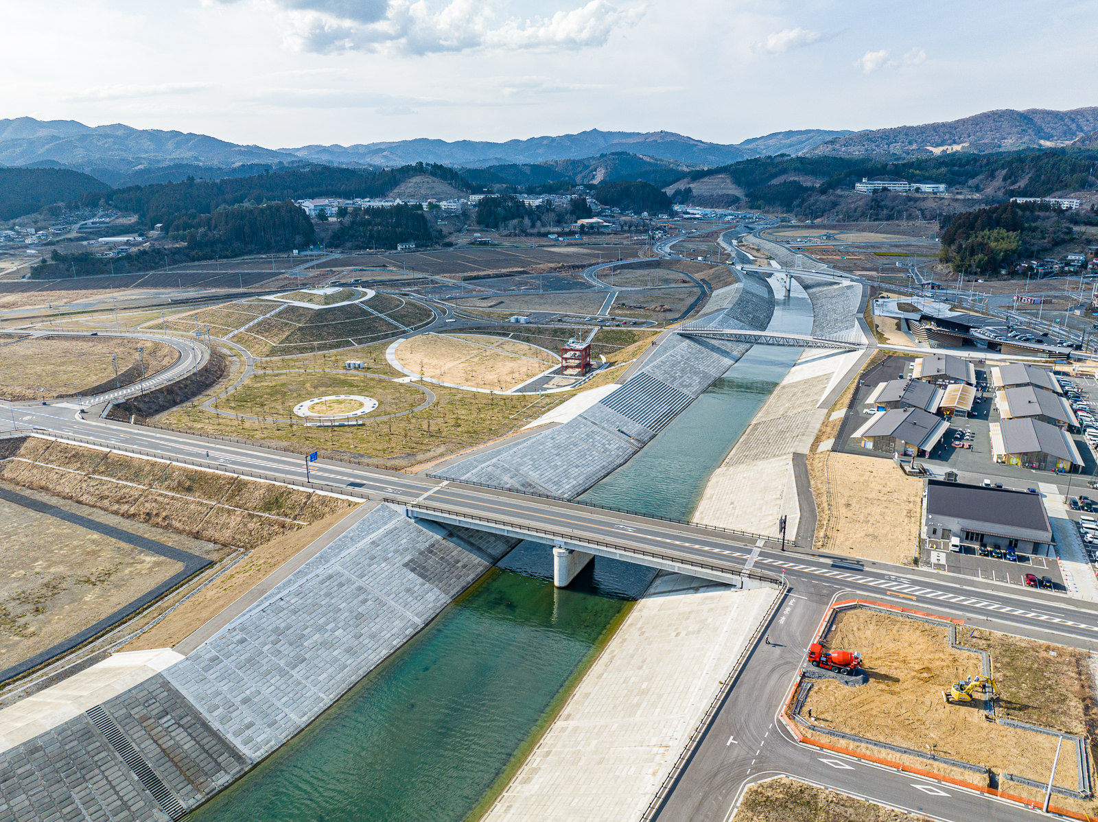整備された川の両岸に道路と土地が整然と配置され、中央にある緑地には複数の円形の植栽スペースが見え、赤い建物も一つ立っており、背景には山が広がる復興した住宅予定地と公園の昼間の風景を高台から定点カメラで撮影した写真