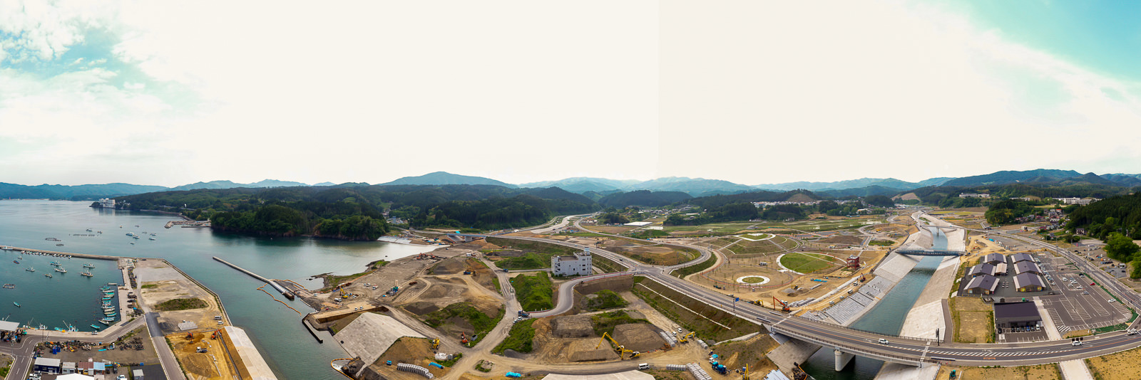 開発または再建中の沿岸地域を広く見渡し、複数の工事現場や道路、インフラ施設が点在し、岸辺には船が停泊し、背景に丘と山々が広がる晴れた空の下の写真