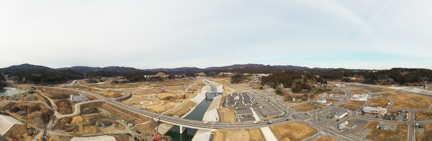 広い範囲にわたる工事現場が広がり、川に架かる橋や複数の道路、点在する建物が見え、周囲には自然の風景も広がっている写真