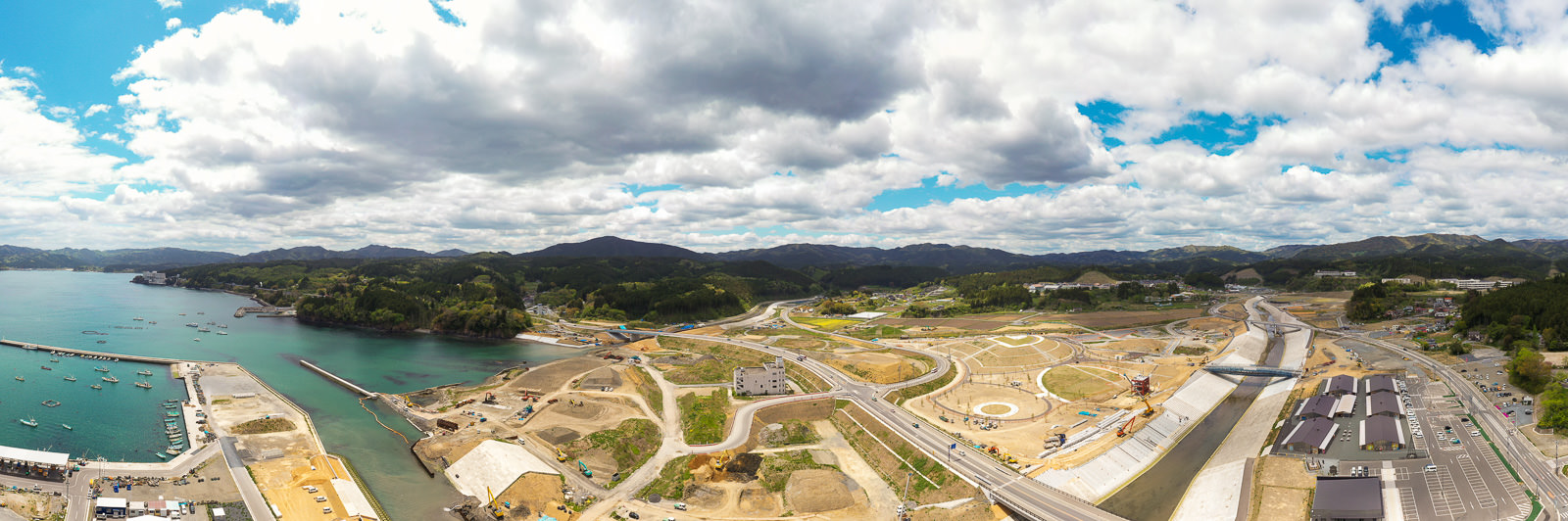 海岸沿いの広い範囲で道路や建物の建設が進められており、背景には緑の山々と澄んだ青空が広がっている工事現場の写真
