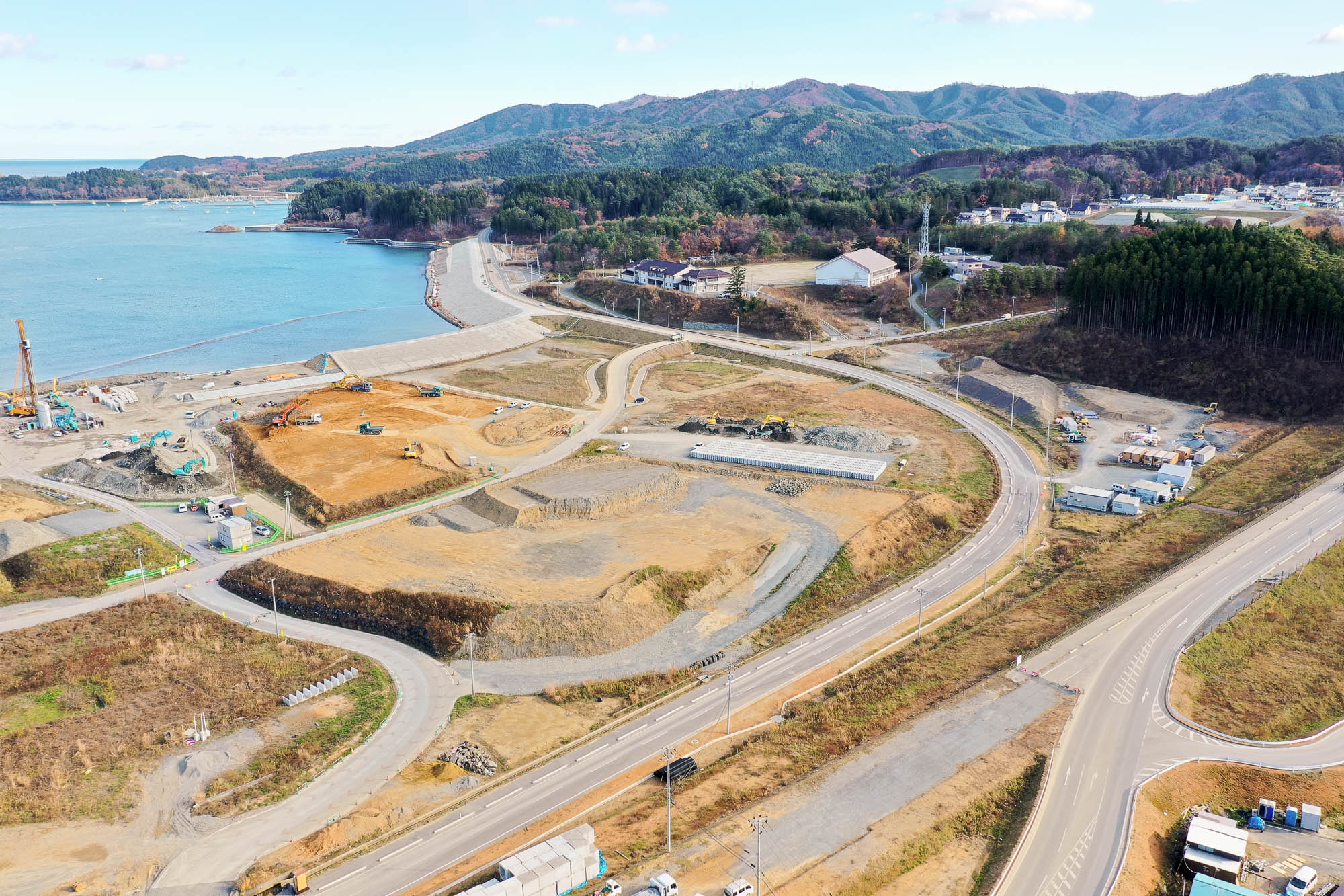 工事中の復興エリアが広がり、防潮堤が湾を縁取るように続き、中央には重機の動く造成地がある写真