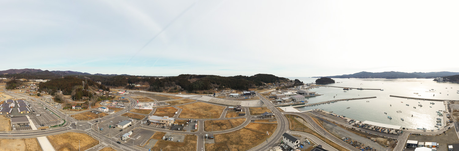 晴れた空の下、右側に船が停泊する水辺が広がり、町には建物や道路、駐車場が点在し、背景には丘や森林が見える海沿いの町の写真