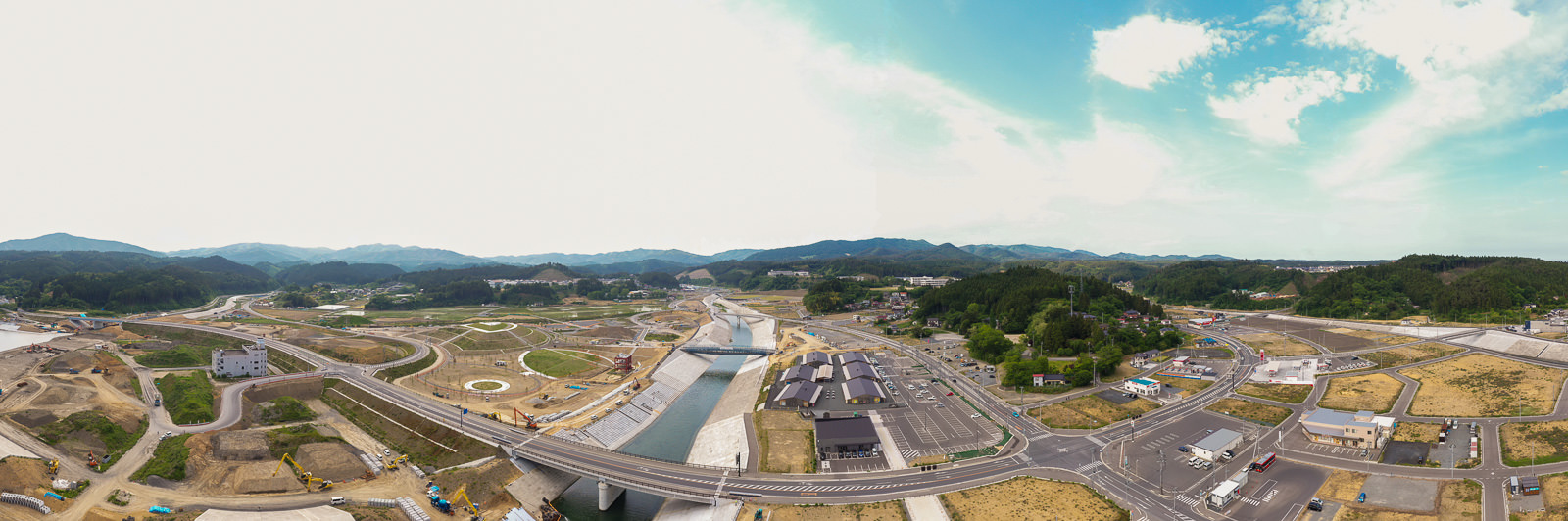 中央を川が流れ、複数の橋がそれを横断し、周囲には道路や建物、広い空き地が広がり、背景には山々が連なり、空には雲が浮かぶ都市と自然が融合した地域の写真
